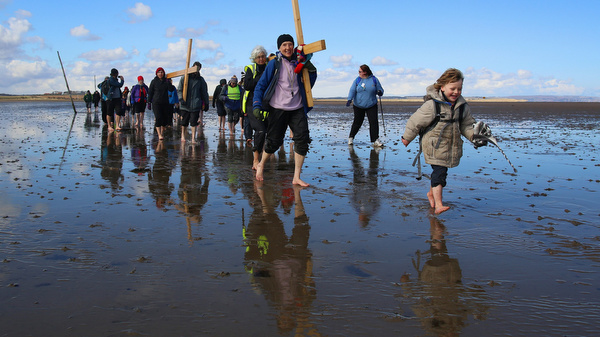 Pilgrims walk across tidal causeway while carrying crosses during pilgrimage in northern England