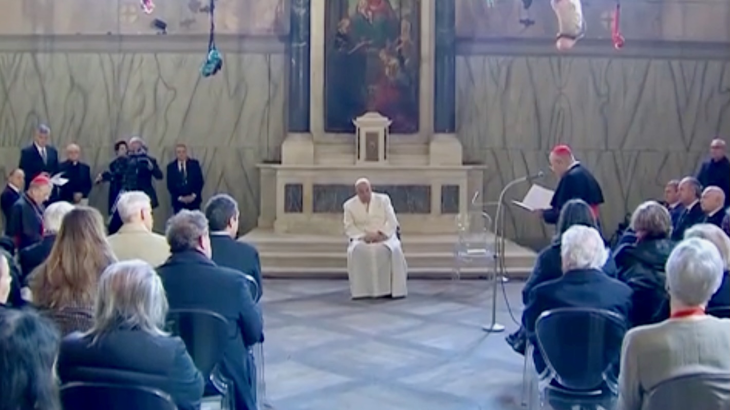 In a full room, Pope Francis is seated centre, Cardinal de Mendonca speaking to him standing at a microphone to the right.