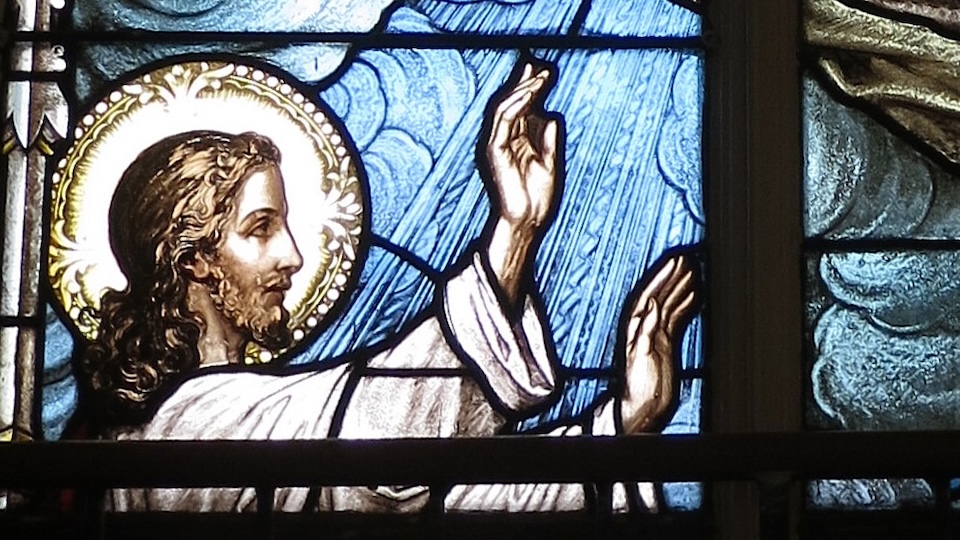 Stained glass of Jesus with a gold halo and serene expression, hands stretched forward.