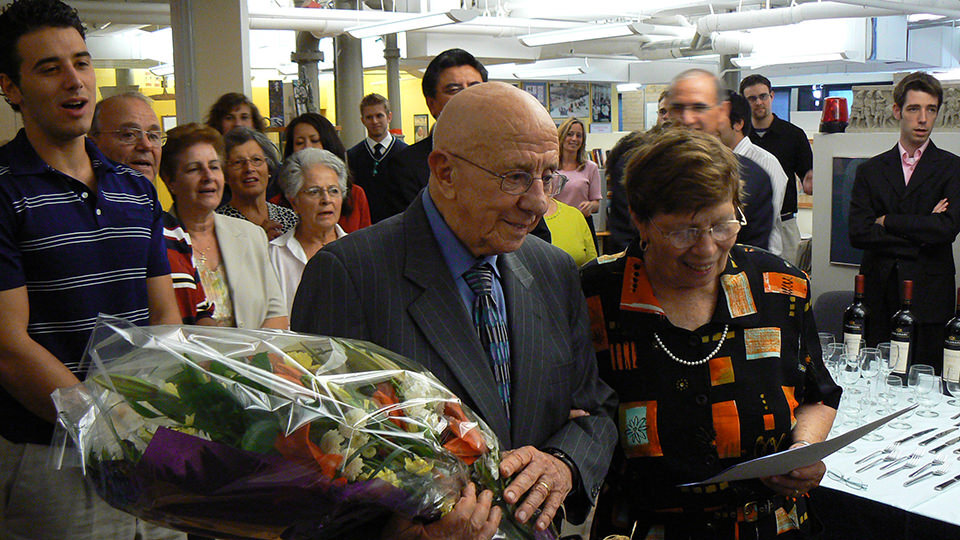A moderate crowd of people stand behind a middle-aged couple, the man holding a bouquet of colourful flowers.